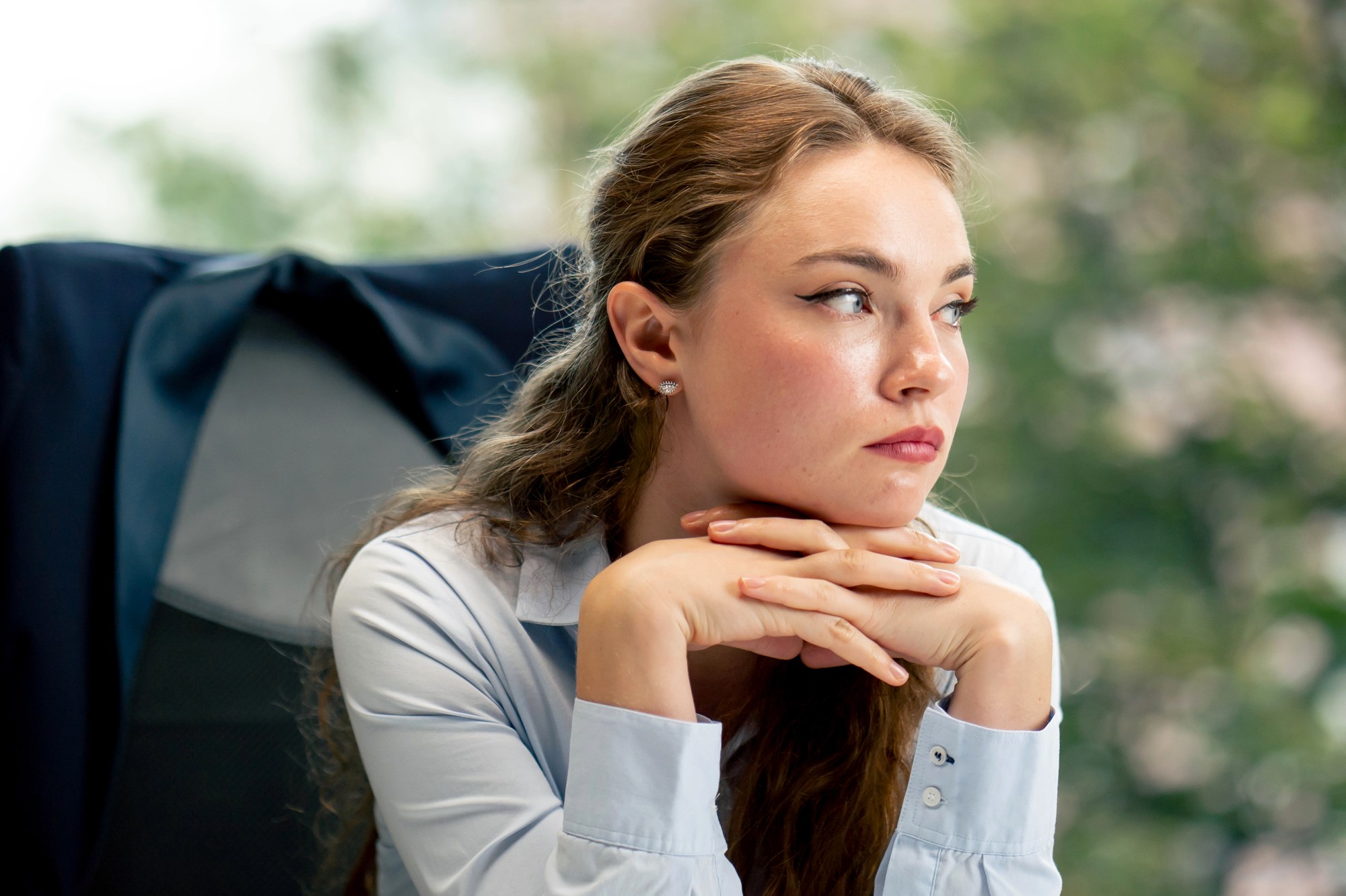 A woman with long brown hair sits at a desk, wearing a light blue blouse. She leans her chin on her hands and gazes thoughtfully to the side. A dark jacket is draped over the chair, and a blurred background of greenery is visible.