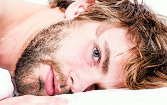 Close-up of a person lying down with their head resting on a white surface. They have light brown hair, a scruffy beard, and blue eyes, looking directly at the camera. The lighting is soft, highlighting their facial features.