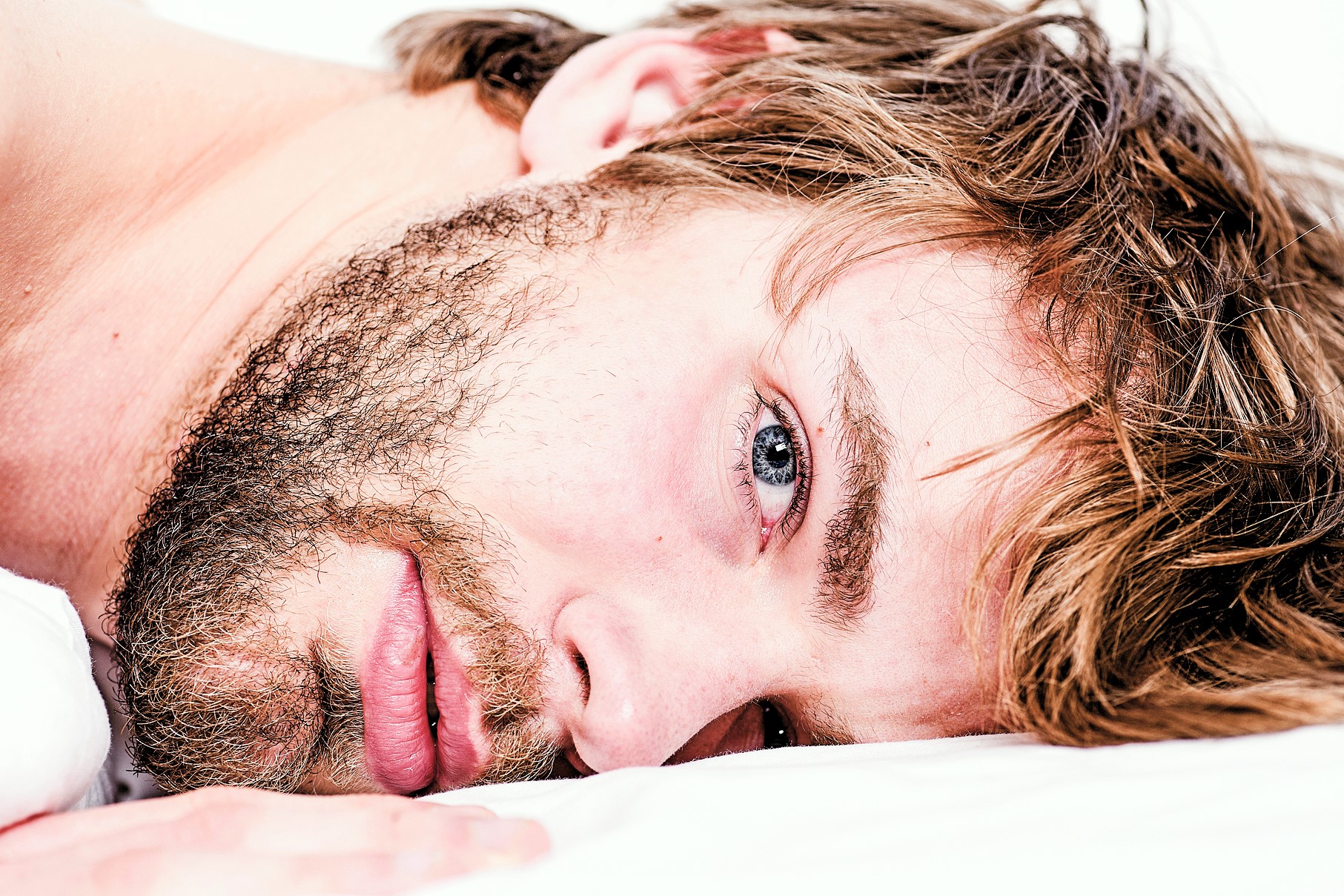 Close-up of a person lying down with their head resting on a white surface. They have light brown hair, a scruffy beard, and blue eyes, looking directly at the camera. The lighting is soft, highlighting their facial features.