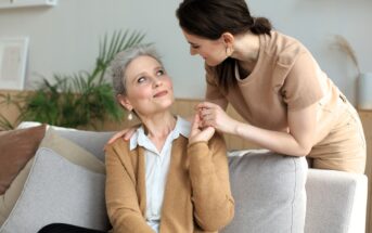 A young woman leans over a couch, smiling warmly at an older woman who is seated, looking up with a smile. They hold hands affectionately. The room is decorated with plants and neutral tones.