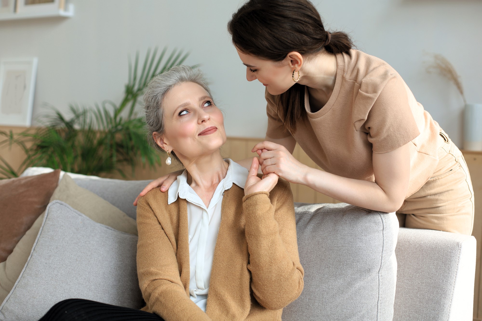 A young woman leans over a couch, smiling warmly at an older woman who is seated, looking up with a smile. They hold hands affectionately. The room is decorated with plants and neutral tones.