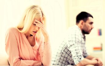 A woman with blonde hair sits in a pink blouse, resting her forehead on her hand, appearing distressed. In the background, a man with dark hair and a beard, wearing a checkered shirt, sits turned away, looking upset.