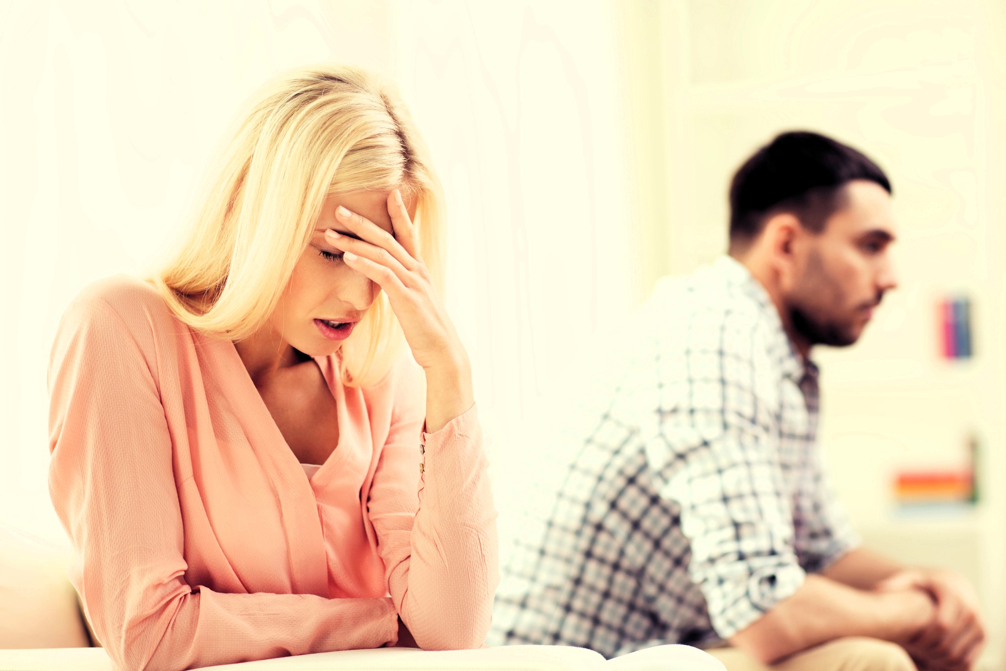 A woman with blonde hair sits in a pink blouse, resting her forehead on her hand, appearing distressed. In the background, a man with dark hair and a beard, wearing a checkered shirt, sits turned away, looking upset.