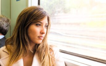 A woman with long brown hair looks contemplatively out a train window. She is wearing a light pink coat. Sunlight illuminates her face, and blurry trees and tracks are visible outside. Another person sits nearby, facing away.