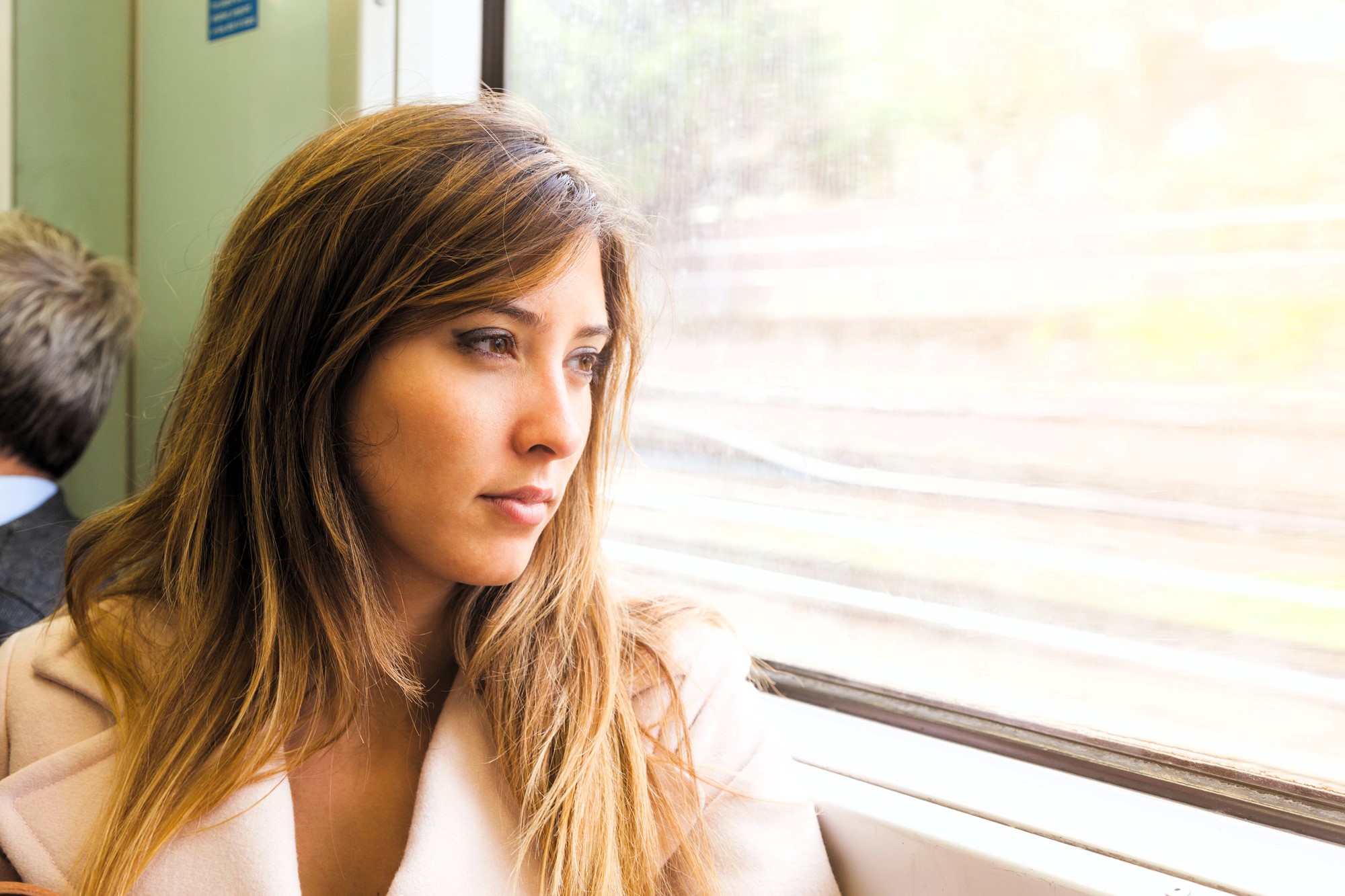 A woman with long brown hair looks contemplatively out a train window. She is wearing a light pink coat. Sunlight illuminates her face, and blurry trees and tracks are visible outside. Another person sits nearby, facing away.