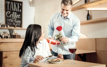 A man in a light blue shirt presents a red rose and a gift to a woman sitting at a table. The woman, smiling, looks up at him. A glass of red wine and a plate of food are in front of her. A "Take Coffee" sign is visible in the background.