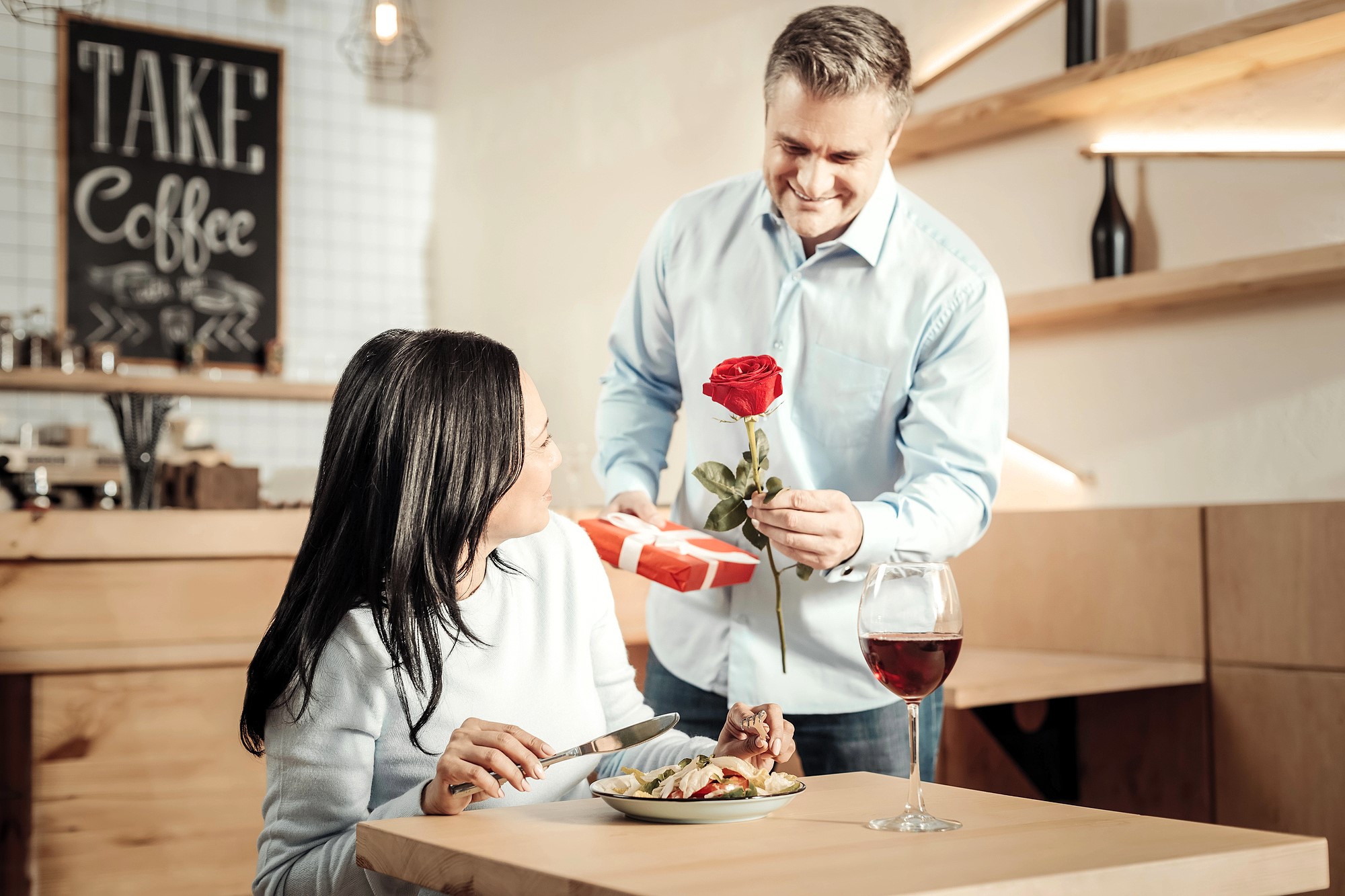 A man in a light blue shirt presents a red rose and a gift to a woman sitting at a table. The woman, smiling, looks up at him. A glass of red wine and a plate of food are in front of her. A "Take Coffee" sign is visible in the background.