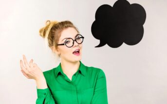 A woman with glasses and a messy bun is wearing a green shirt and gesturing with her hand. She stands next to a large black speech bubble on a gray background. Her expression is surprised or intrigued.