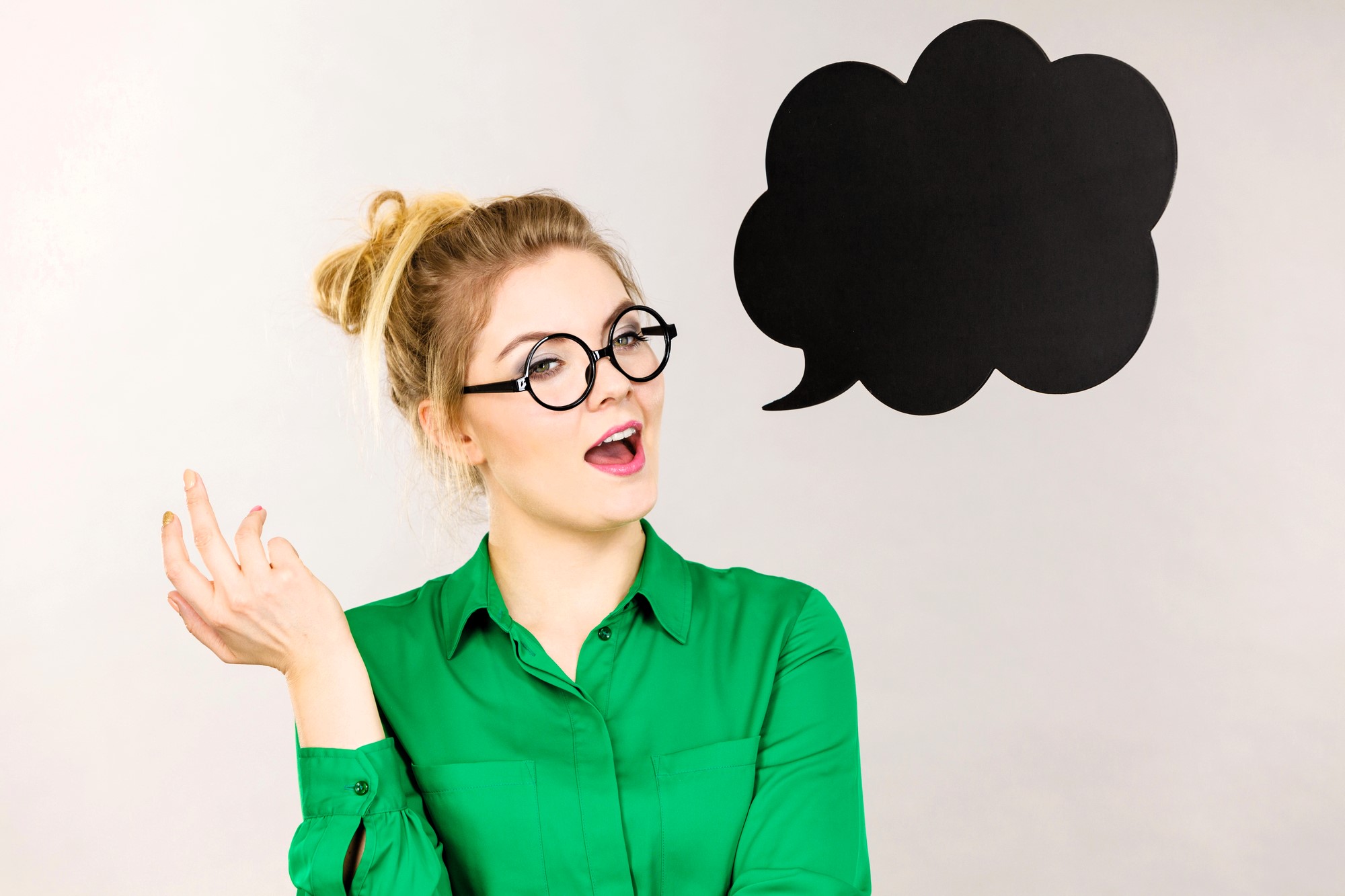 A woman with glasses and a messy bun is wearing a green shirt and gesturing with her hand. She stands next to a large black speech bubble on a gray background. Her expression is surprised or intrigued.