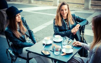 Three women are sitting at an outdoor café table, enjoying coffee. They are dressed in stylish casual outfits with jackets. The table is set with cups, glasses, and a phone. The setting appears relaxed and social.