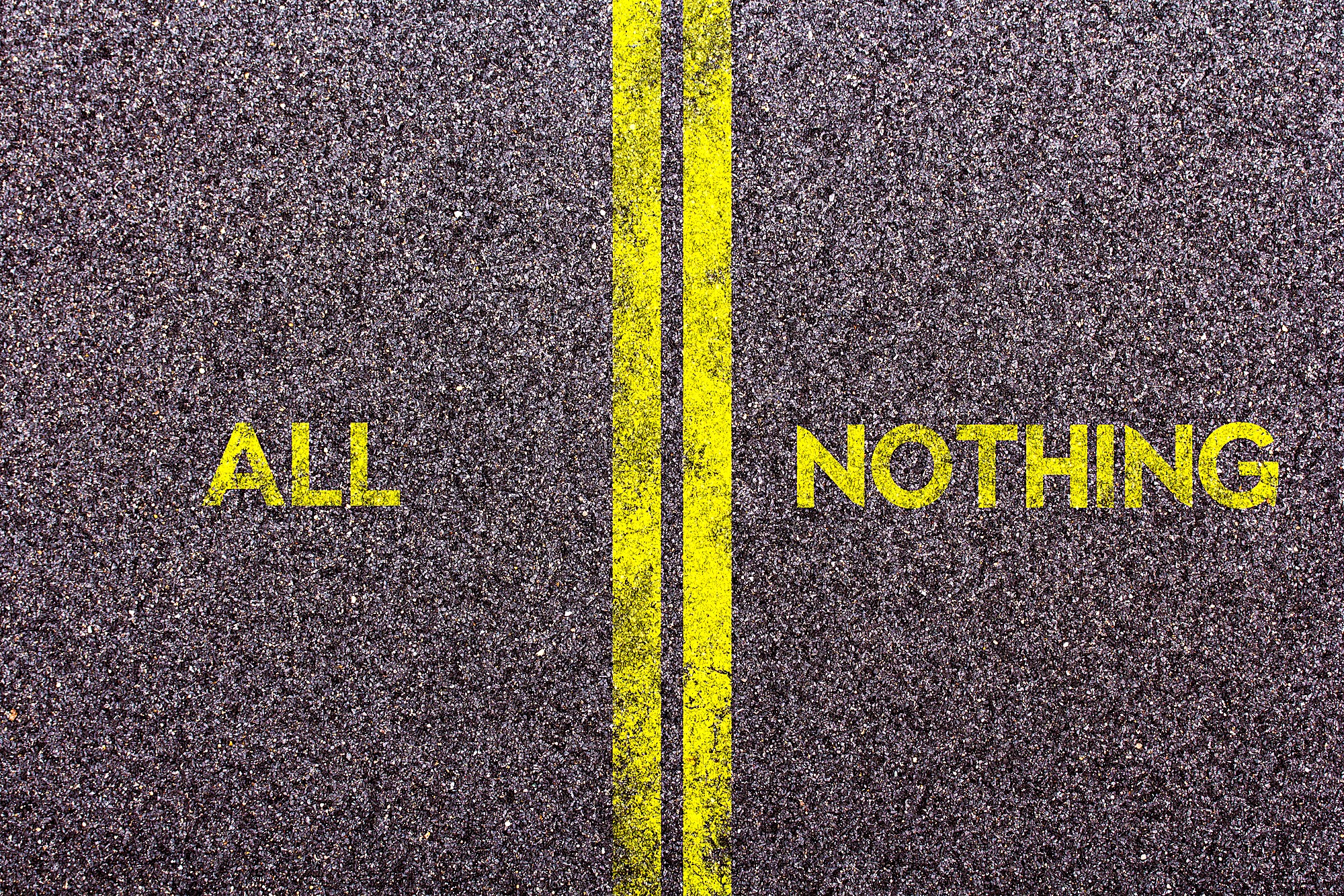 A road with a double yellow line in the middle. The word "ALL" is painted on one side of the line, and "NOTHING" is on the other, both in yellow. The texture of the asphalt is visible.