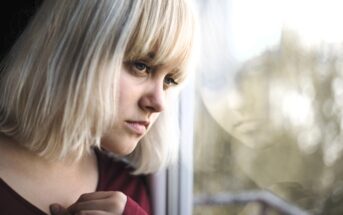 A woman with blonde hair gazes thoughtfully out of a window. Her reflection is visible in the glass. She wears a maroon top and appears deep in contemplation. The background is softly blurred, suggesting an outdoor setting.