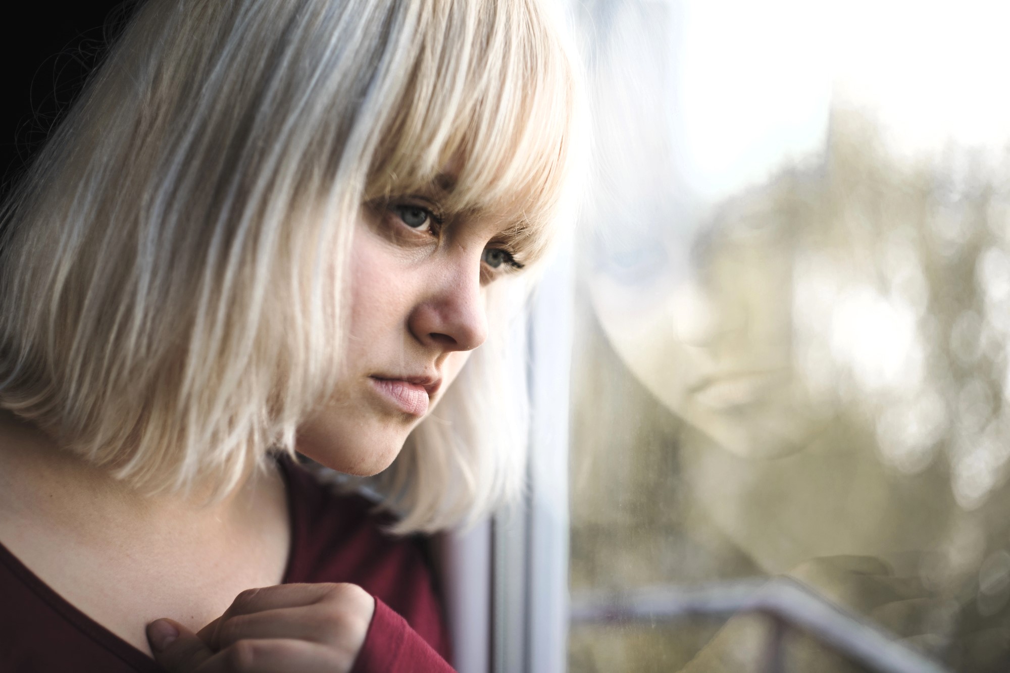 A woman with blonde hair gazes thoughtfully out of a window. Her reflection is visible in the glass. She wears a maroon top and appears deep in contemplation. The background is softly blurred, suggesting an outdoor setting.