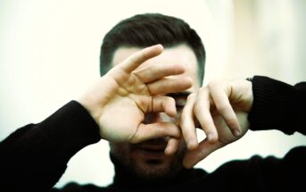 A person wearing a black turtleneck covers their face with their hands, partially obscuring their features. The background is softly blurred, creating a gentle focus on the hands and face.