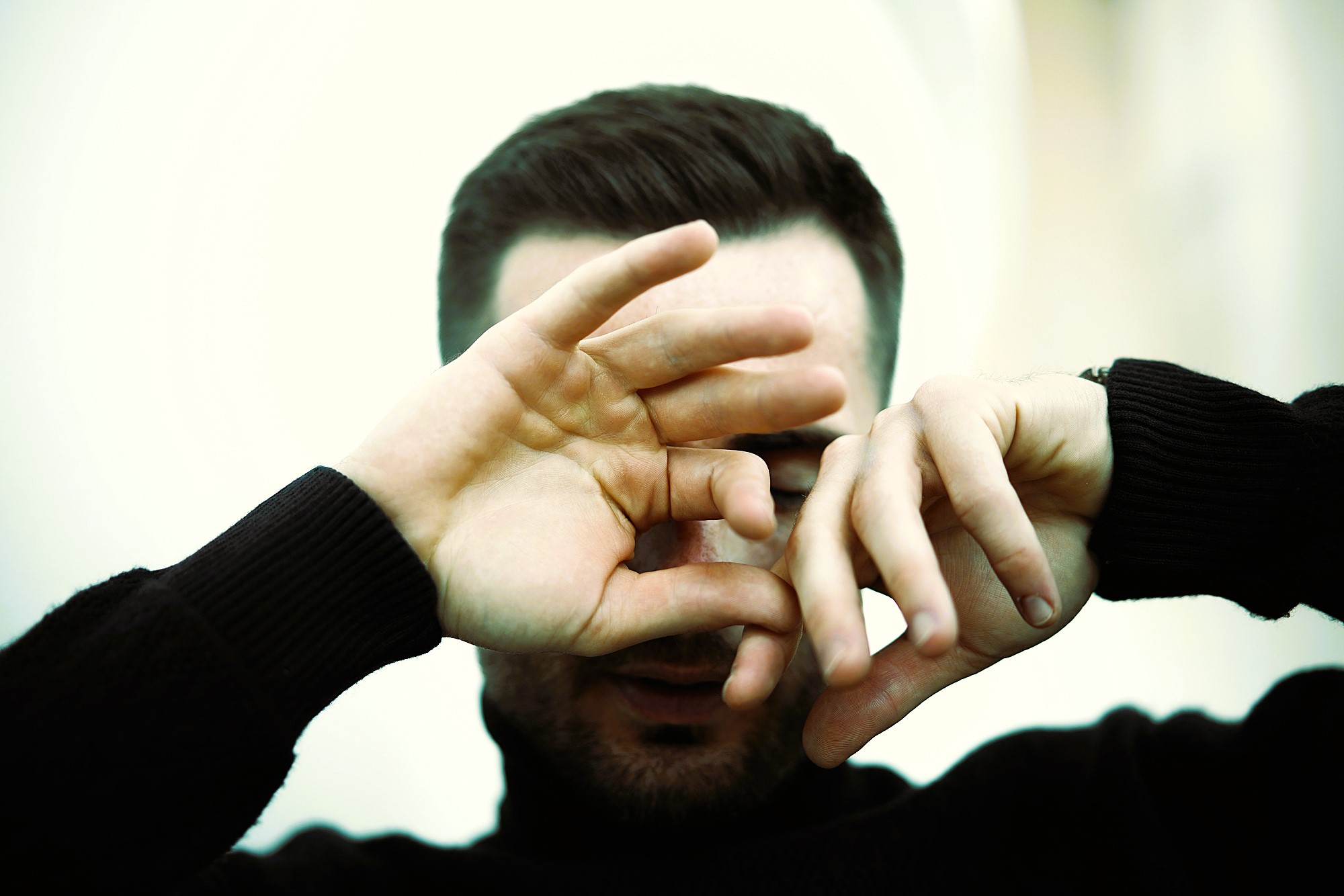 A person wearing a black turtleneck covers their face with their hands, partially obscuring their features. The background is softly blurred, creating a gentle focus on the hands and face.