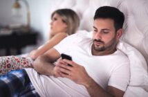 A man in a white t-shirt and blue plaid pants lies on a bed, looking at his smartphone. A woman with blonde hair, wearing a white top and floral pants, is lying beside him, facing away. The room has a soft, neutral color palette.