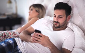 A man in a white t-shirt and blue plaid pants lies on a bed, looking at his smartphone. A woman with blonde hair, wearing a white top and floral pants, is lying beside him, facing away. The room has a soft, neutral color palette.