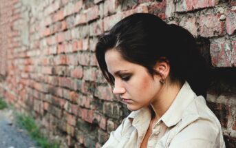 A woman with dark hair tied back sits against a brick wall, looking downward. She is wearing a beige shirt and appears thoughtful or pensive. The brick wall extends into the distance on her left side.