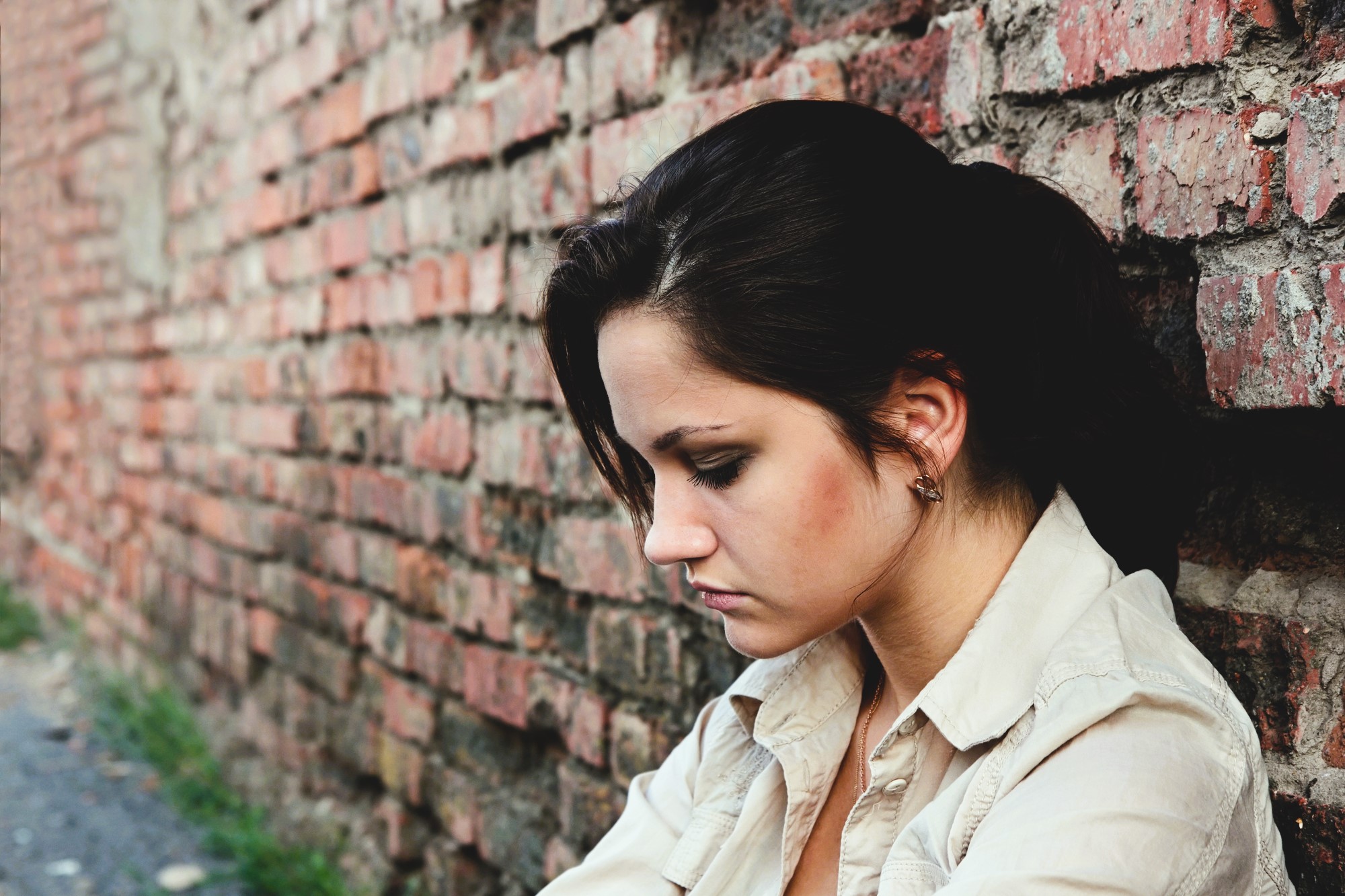 A woman with dark hair tied back sits against a brick wall, looking downward. She is wearing a beige shirt and appears thoughtful or pensive. The brick wall extends into the distance on her left side.