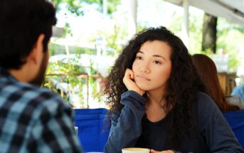 Woman with curly hair, wearing a blue sweater, sits at an outdoor café table, resting her chin on her hand while looking at another person. She holds a coffee cup, and greenery is visible in the background.