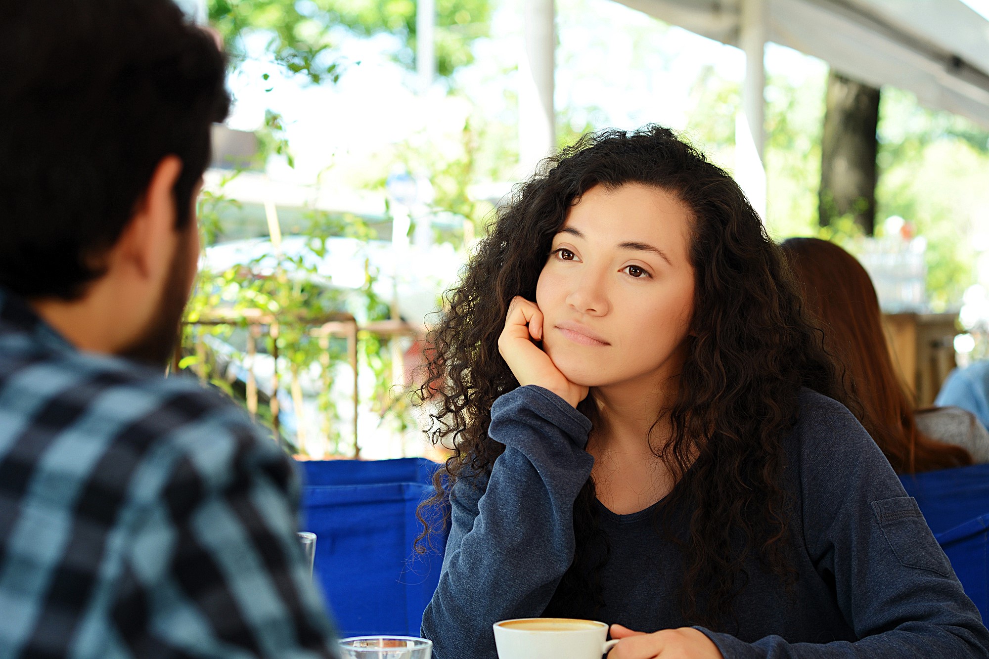 Woman with curly hair, wearing a blue sweater, sits at an outdoor café table, resting her chin on her hand while looking at another person. She holds a coffee cup, and greenery is visible in the background.