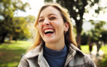 A woman with long hair laughs heartily outdoors in a park. She wears a blue turtleneck and a jacket, with trees and blurred people in the background. The atmosphere is joyful and relaxed.