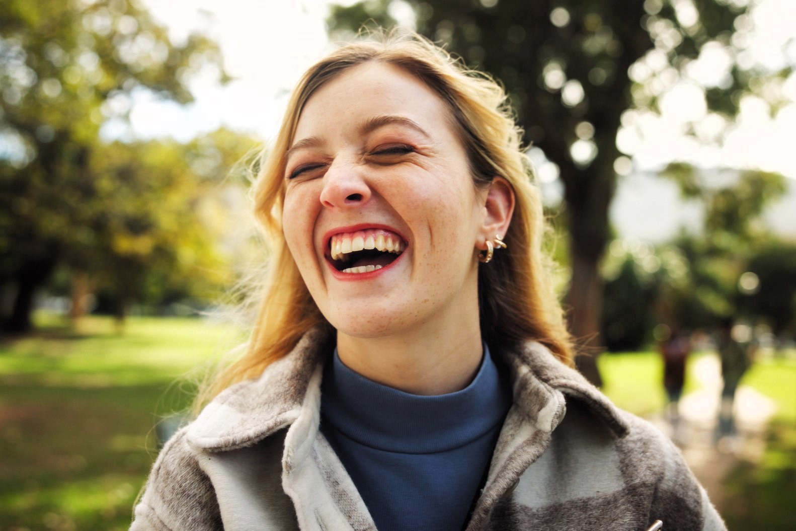 A woman with long hair laughs heartily outdoors in a park. She wears a blue turtleneck and a jacket, with trees and blurred people in the background. The atmosphere is joyful and relaxed.