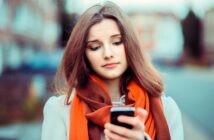 A young woman with long brown hair looks at her phone with a slightly concerned expression. She is wearing an orange scarf and a white coat, standing outdoors with blurred buildings in the background.