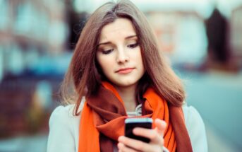 A young woman with long brown hair looks at her phone with a slightly concerned expression. She is wearing an orange scarf and a white coat, standing outdoors with blurred buildings in the background.