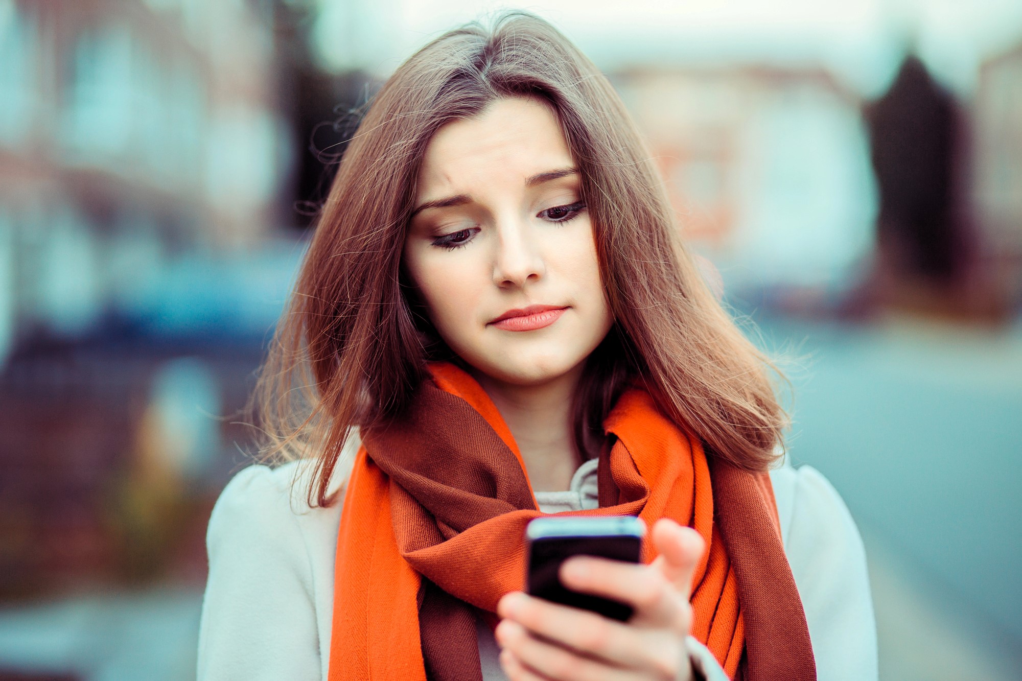 A young woman with long brown hair looks at her phone with a slightly concerned expression. She is wearing an orange scarf and a white coat, standing outdoors with blurred buildings in the background.