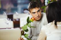 A young man with short brown hair wearing a light gray sweater holds a yellow rose with red edges, looking intently at a person with dark hair, whose back is facing the camera. They are seated in a softly lit restaurant.