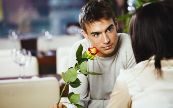 A young man with short brown hair wearing a light gray sweater holds a yellow rose with red edges, looking intently at a person with dark hair, whose back is facing the camera. They are seated in a softly lit restaurant.