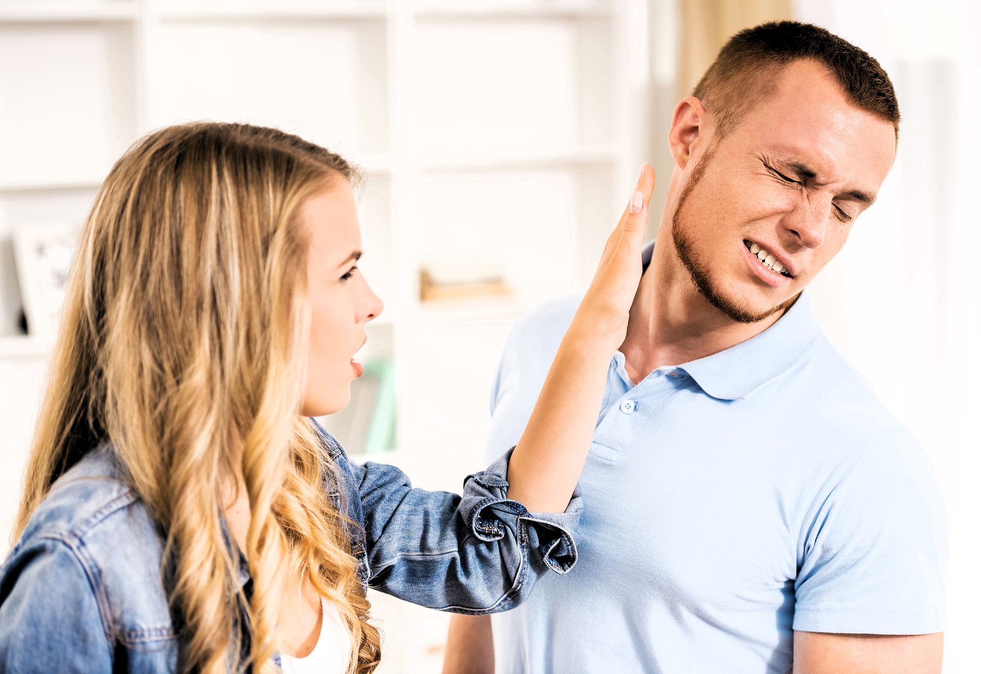 A woman with long blonde hair appears to be in the middle of slapping a man who is wincing in pain. Both are dressed in casual clothing, and the background shows a blurred indoor setting.