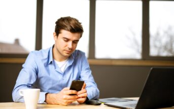 A man in a blue shirt sits at a wooden table, holding a smartphone. A white coffee mug, a black computer mouse, and an open laptop are on the table. Large windows in the background let in natural light.