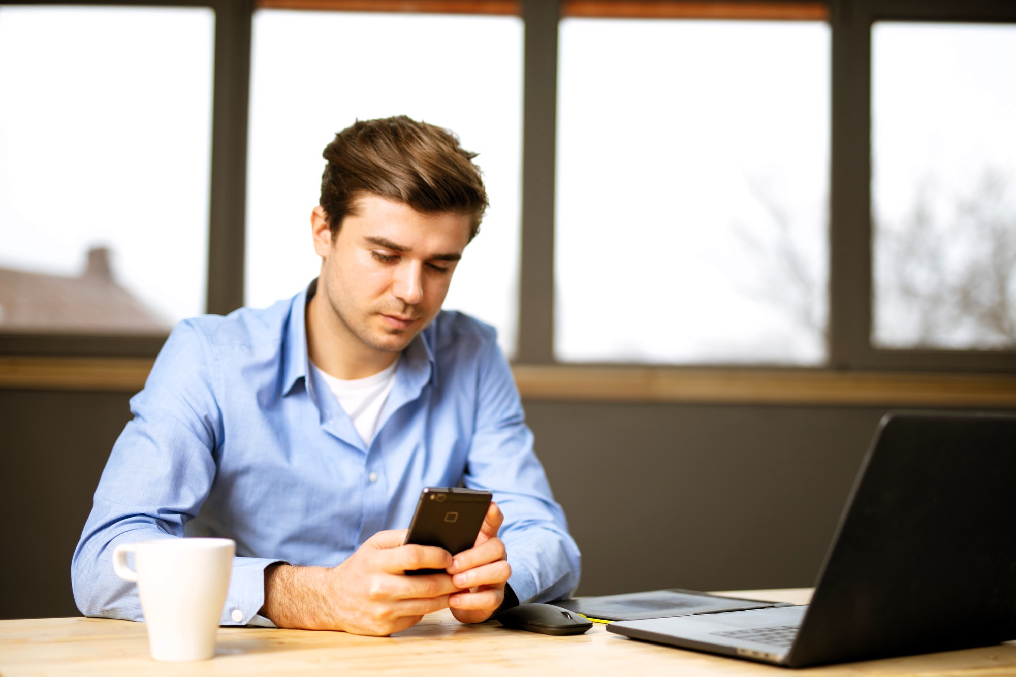 A man in a blue shirt sits at a wooden table, holding a smartphone. A white coffee mug, a black computer mouse, and an open laptop are on the table. Large windows in the background let in natural light.