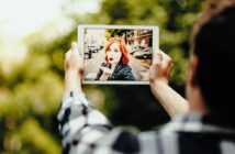 A person holds a tablet in both hands, capturing an image of a woman with red hair smiling and posing in an urban setting. Trees and a street with parked cars are visible in the background.