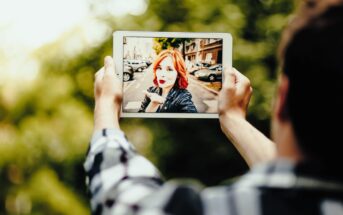 A person holds a tablet in both hands, capturing an image of a woman with red hair smiling and posing in an urban setting. Trees and a street with parked cars are visible in the background.