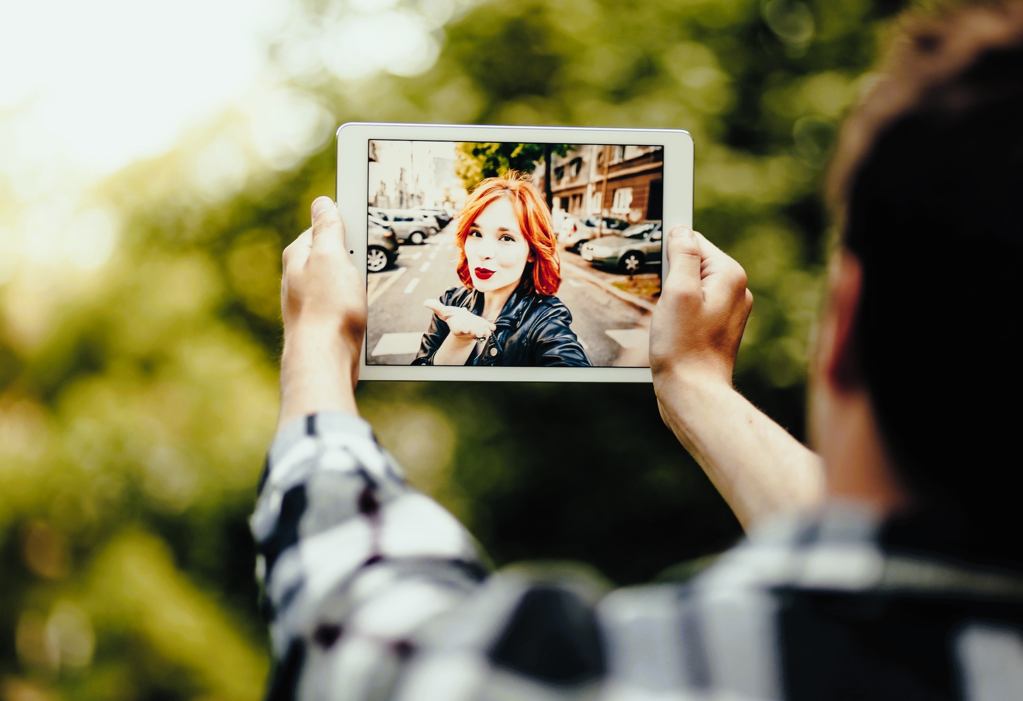 A person holds a tablet in both hands, capturing an image of a woman with red hair smiling and posing in an urban setting. Trees and a street with parked cars are visible in the background.