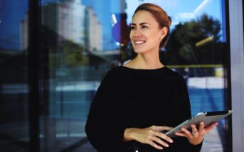 A woman in a black top stands outside holding a tablet. She is smiling and looking to the side. The background shows glass doors reflecting buildings and trees in daylight.