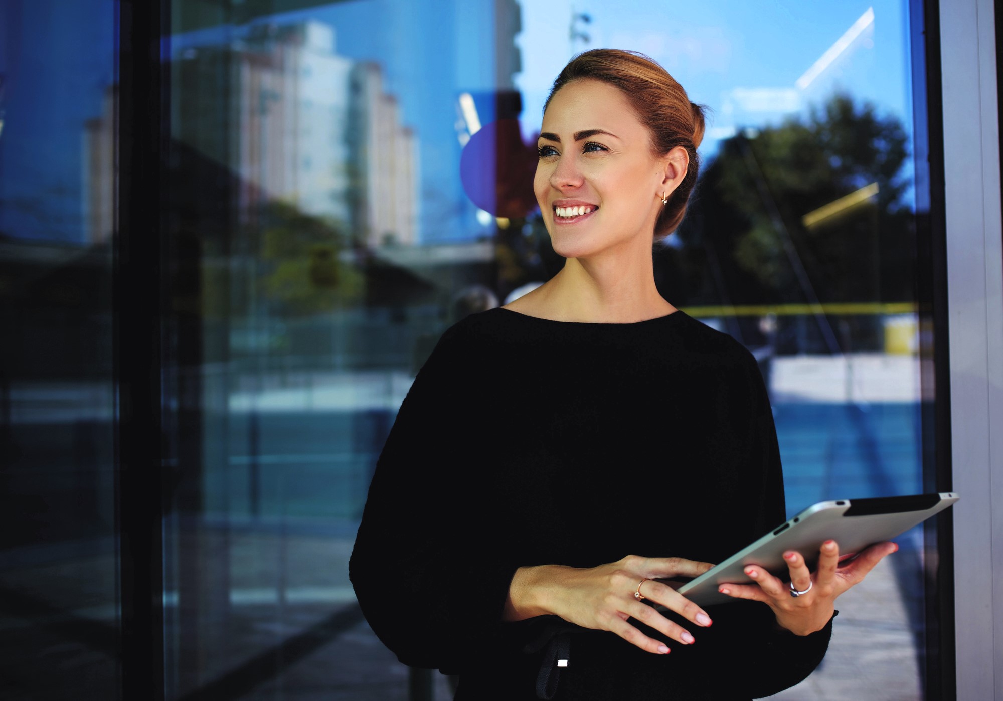 A woman in a black top stands outside holding a tablet. She is smiling and looking to the side. The background shows glass doors reflecting buildings and trees in daylight.