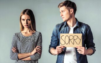 A man in a denim shirt holds a cardboard sign that says "SORRY," looking at a woman in a striped shirt who appears upset with crossed arms. They stand against a plain gray background.