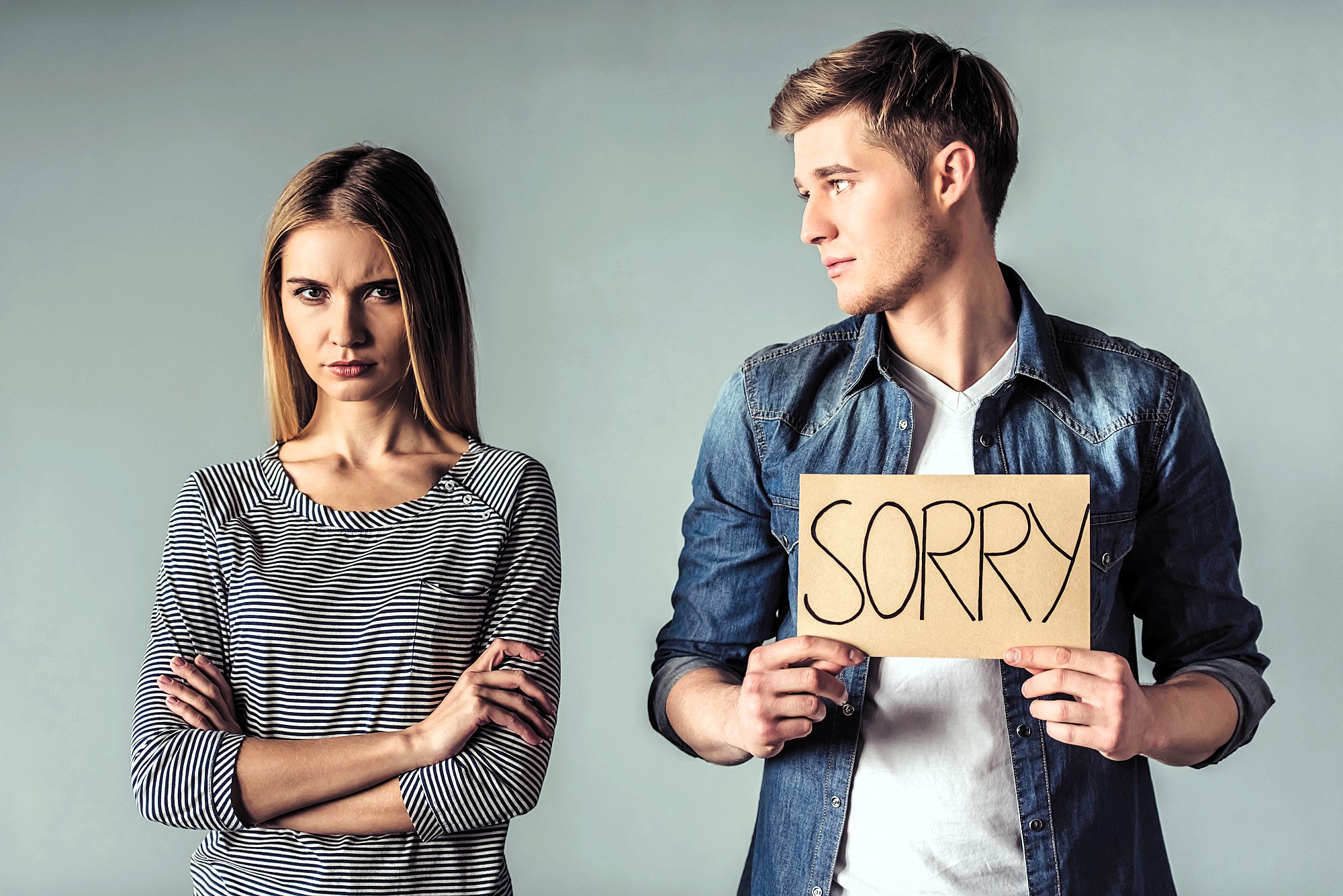 A man in a denim shirt holds a cardboard sign that says "SORRY," looking at a woman in a striped shirt who appears upset with crossed arms. They stand against a plain gray background.