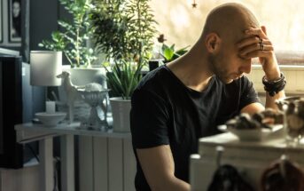 A man with a shaved head sits at a table in a dimly lit room, resting his forehead on his hand. He wears a black t-shirt and rings. In the background are plants, a lamp, and various decorative items. Natural light filters in from a window.