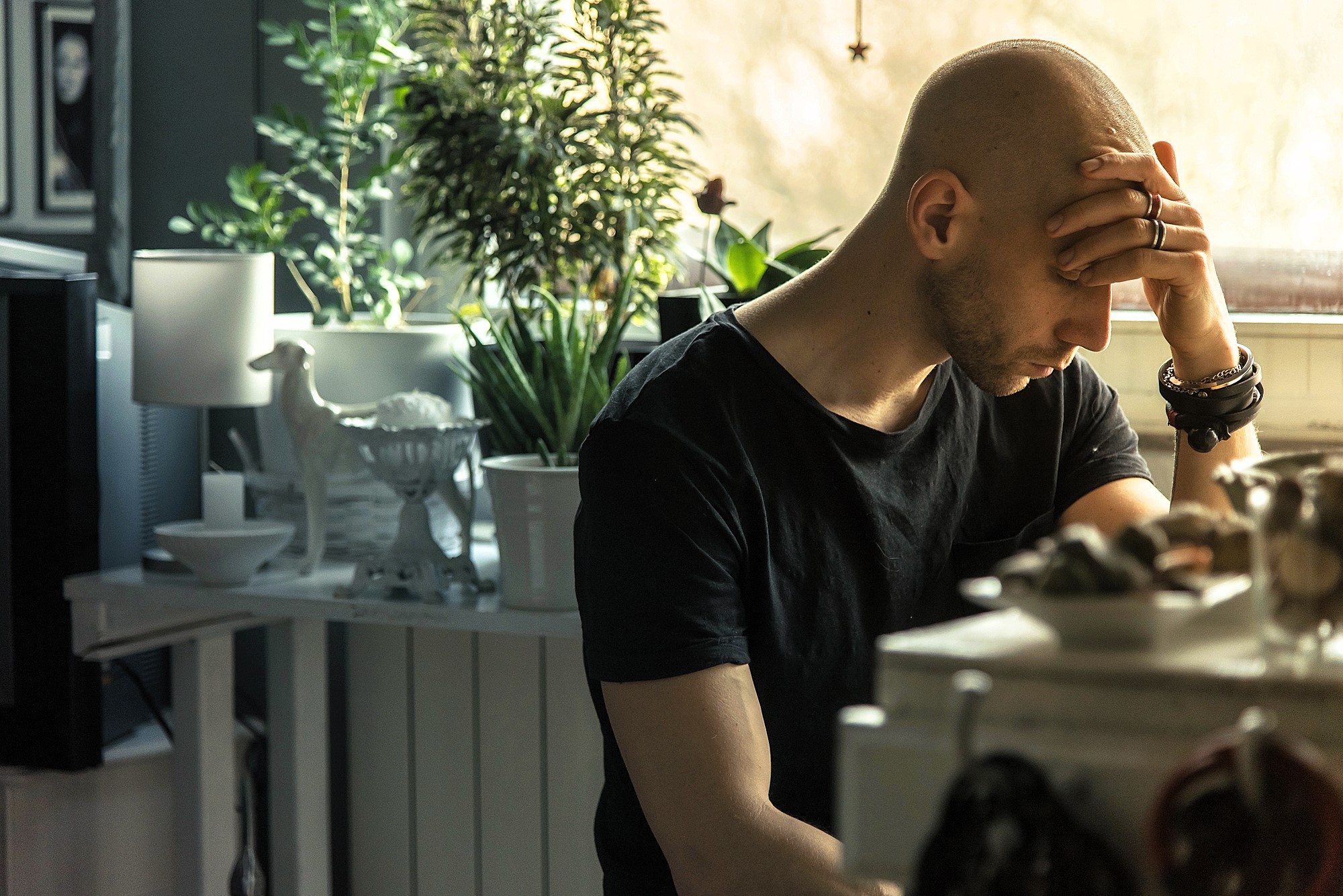 A man with a shaved head sits at a table in a dimly lit room, resting his forehead on his hand. He wears a black t-shirt and rings. In the background are plants, a lamp, and various decorative items. Natural light filters in from a window.