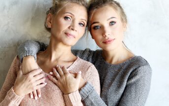 Two women with light hair stand close together against a textured, light-colored wall. They wear knitted sweaters, one in pink and the other in gray, looking calmly at the camera with gentle expressions and natural makeup.