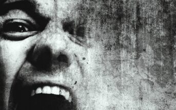 A close-up of a face in black and white, showing one eye and an open mouth with visible teeth, giving an expression of intense emotion. The image has a textured, gritty overlay, adding to the dramatic effect.