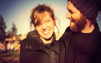 A woman and a man standing close, both smiling warmly. The woman is looking at the camera, while the man looks at her. They are wearing winter clothing, and the background shows a blurred outdoor scene with trees.