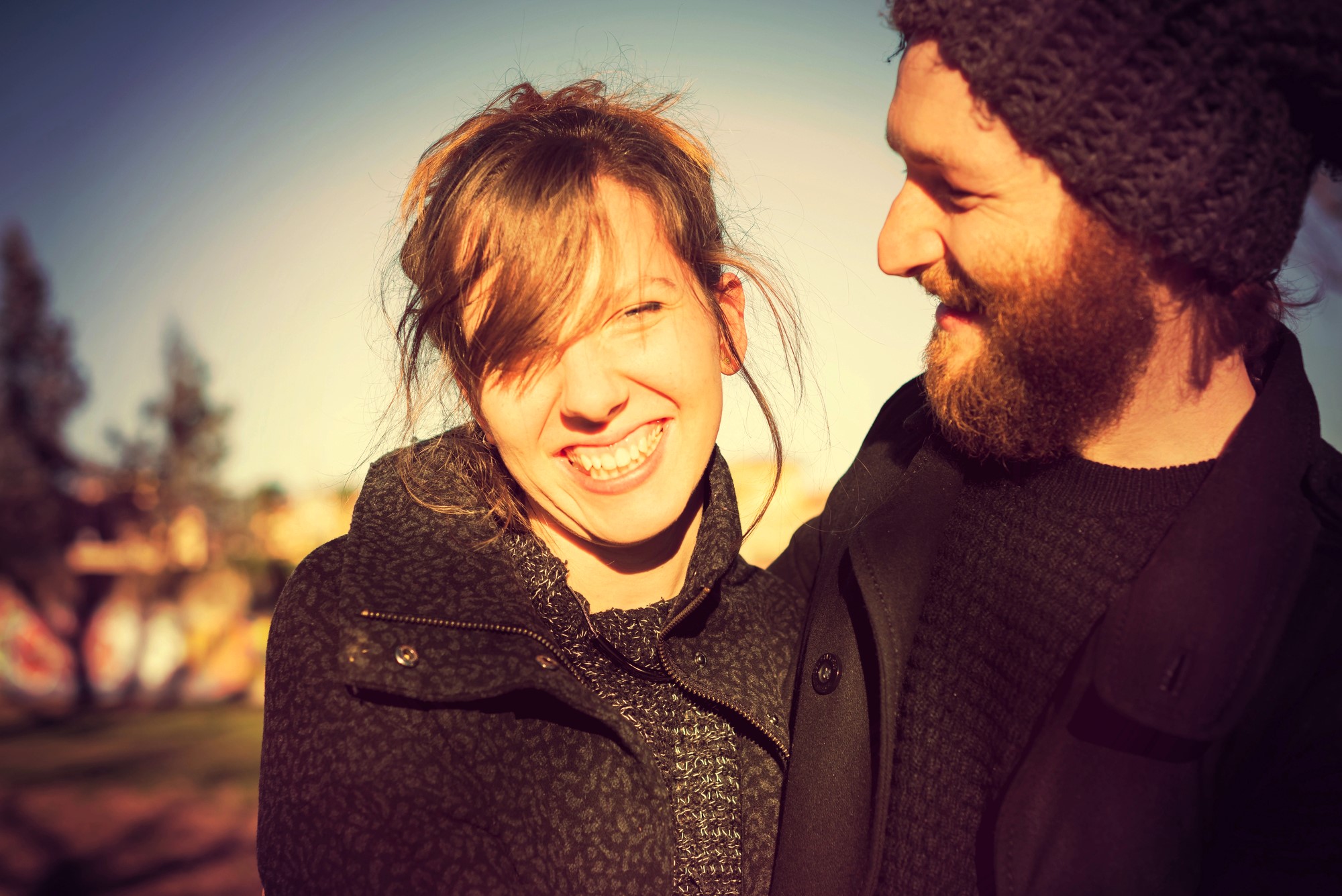 A woman and a man standing close, both smiling warmly. The woman is looking at the camera, while the man looks at her. They are wearing winter clothing, and the background shows a blurred outdoor scene with trees.