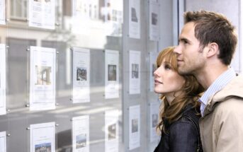 A man and woman are standing side by side, looking at property listings displayed in a window. The man is wearing a jacket and the woman has a denim jacket. They appear to be interested in the real estate advertisements in front of them.