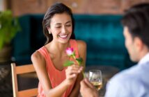 A woman smiles while receiving a pink rose from a person sitting across from her. She is holding a glass of white wine. They are seated at a table in a warmly lit setting with a green velvet sofa in the background.